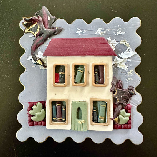 Decorative house ornament with book-themed windows on a textured background with flying dragons. Wall art or brooch available.